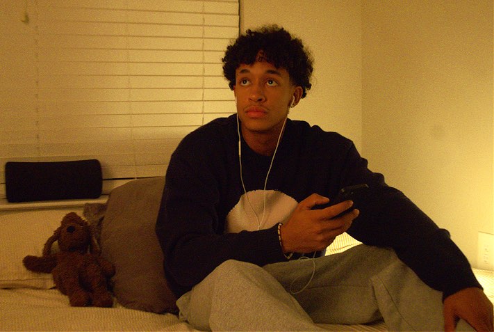 Seated on a bed at home in the Merino Knit, headphones in.