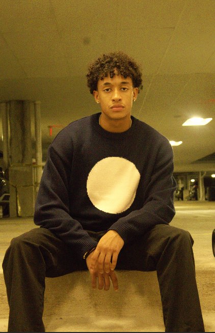 The Merino Knit worn full-length with black trousers, seated in a parking garage.